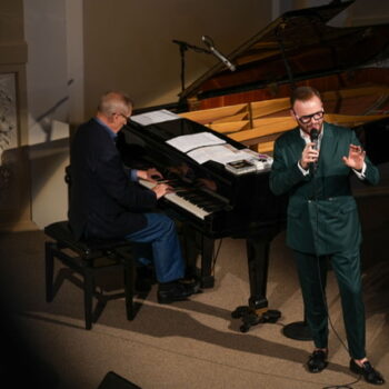 Emil Viklický seated at a piano and Jan Smigmator with a microphone in hand.