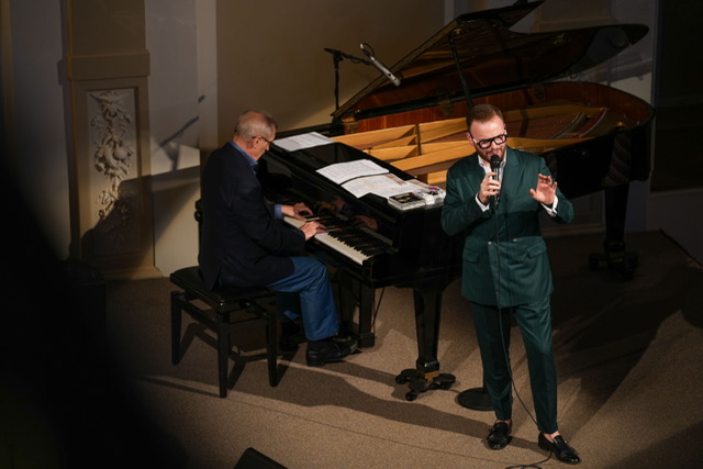 Emil Viklický seated at a piano and Jan Smigmator with a microphone in hand.